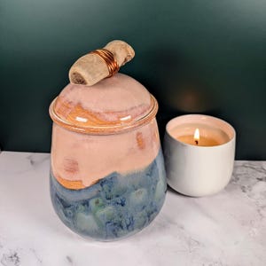 May include: A handcrafted ceramic jar with a lid, displaying a colour gradient from pink to blue. The lid is topped with a small, light-coloured stone wrapped in copper wire. A lit white candle sits nearby, creating a cosy ambience.