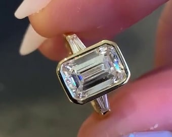 May include: A gold ring featuring a large, rectangular emerald-cut diamond set in a bezel setting. The diamond is flanked by smaller, baguette-cut diamonds on each side.