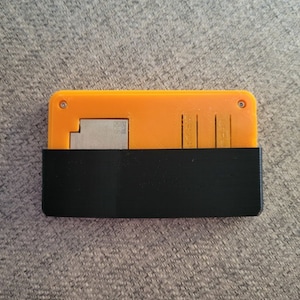 Pode incluir: Uma pequena caixa de plástico laranja e preta com um inserto de metal. A caixa tem dois pequenos furos na parte superior.