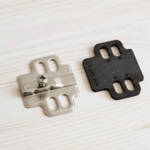 Op de afbeelding: Een zilveren kastdeur scharnier met een schroef en een zwarte plastic sjabloon met de tekst "WAR GAP KIT".