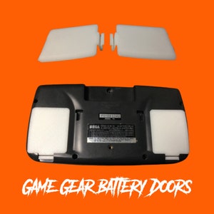 Könnte beinhalten: Zwei weiße Plastik-Batteriedeckel für eine schwarze Game Gear Handheld-Videospielkonsole. Die Konsole trägt den Text "SEGA" und "P31063342".