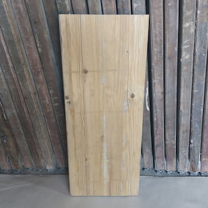 May include: A rectangular, light-colored wooden plank with visible grain and knots. The plank is positioned vertically against a backdrop of weathered, dark-toned wooden planks. The wood has a natural, rustic appearance.