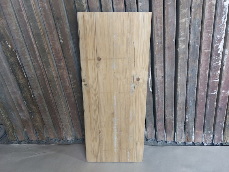 May include: A rectangular, light-colored wooden plank with visible grain and knots. The plank is positioned vertically against a backdrop of weathered, dark-toned wooden planks. The wood has a natural, rustic appearance.