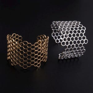 May include: Two cuff bracelets featuring an openwork honeycomb pattern. One bracelet is finished in a metallic bronze tone, while the other is finished in a metallic silver tone. Both pieces have a wide, circular band design with open ends.
