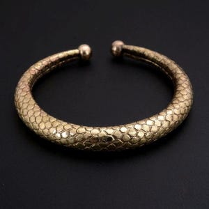 Puede incluir: Una pulsera tipo brazalete de metal en tono dorado con un patrón texturizado de escamas hexagonales repetidas en toda su superficie. La pulsera es abierta, con dos pequeños terminales redondeados y lisos en cada extremo de la banda circular.