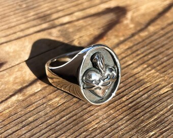 May include: A silver ring with a detailed Sacred Heart design. The ring features a polished band and a raised oval face with the heart symbol. The ring is set against a wooden surface, with sunlight casting shadows.