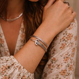 May include: A silver necklace and two silver bracelets are displayed. One bracelet features a decorative sword design. The jewelry is worn on a person's arm, with a floral patterned top in the background.