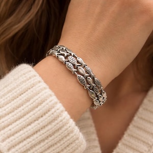 May include: Silver bracelet with a repeating fish pattern. The bracelet has three rows of interconnected fish, creating a detailed accessory. The bracelet is worn on a wrist, highlighting its intricate design and metallic sheen. The bracelet is made of silver.