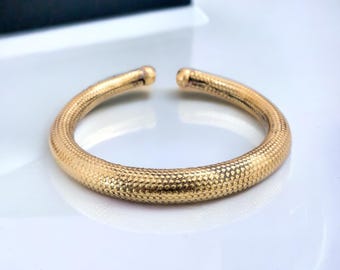 Puede incluir: Un brazalete dorado con una superficie texturizada y punteada. El diseño abierto presenta puntas redondeadas. La joya se exhibe sobre una superficie blanca, con una caja oscura en el fondo.