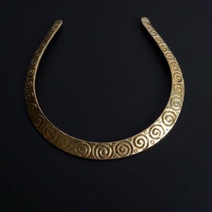 Puede incluir: Un collar tipo torque de metal en tono dorado con un diseño abierto en forma de media luna. La superficie del collar está decorada con un patrón repetitivo de motivos de espirales en relieve y pequeños puntos circulares, creando una apariencia texturizada.