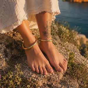 May include: A pair of polished gold-tone metal anklets featuring a smooth, rounded, and continuous circular band design. These minimalist jewelry pieces are worn around the ankles, showcasing a reflective metallic finish that catches the sunlight.