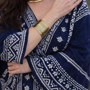 Puede incluir: Un conjunto de brazaletes finos de metal dorado en la muñeca, junto a una prenda azul oscuro con un patrón floral y geométrico blanco. La tela tiene una base azul marino profundo con diseños blancos intrincados en las mangas y el cuerpo.