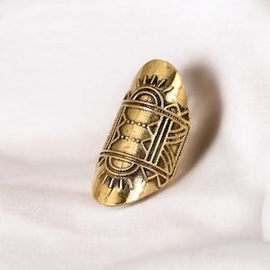 Puede incluir: Un anillo dorado con forma ovalada alargada. El anillo presenta intrincados patrones geométricos, que incluyen líneas, puntos y diseños en forma de sol. El anillo está hecho de metal y tiene un acabado pulido.