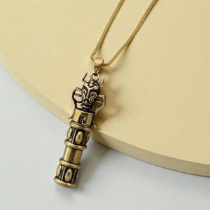 May include: A gold-colored pendant necklace featuring a detailed, cylindrical charm. The charm has an intricate design with a stylized face at the top and a series of circular patterns along its length. The necklace chain is a fine, gold-toned snake chain.