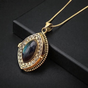 May include: Gold-toned pendant necklace with an oval-shaped locket. The locket features an ornate filigree design and a central, iridescent stone. The necklace is displayed on a black surface, highlighting the jewelry's details.