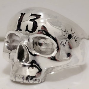 以下が含まれることがあります： 上部に数字の「13」が付いたシルバーのスカルリング。スカルの側面にはひび割れのデザインがあります。このリングはゴシック様式のアクセサリーです。