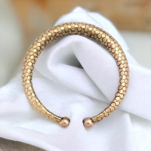May include: A gold-colored cuff bracelet with a textured, scale-like pattern. The open-ended design features rounded terminals. The bracelet is displayed on a white cloth, highlighting its metallic sheen and intricate surface.