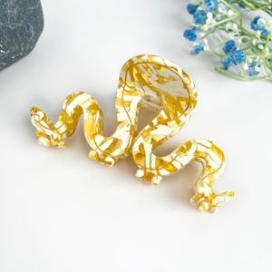 May include: A yellow and white snake-shaped hair clip. The clip is made of plastic and has a glossy finish.