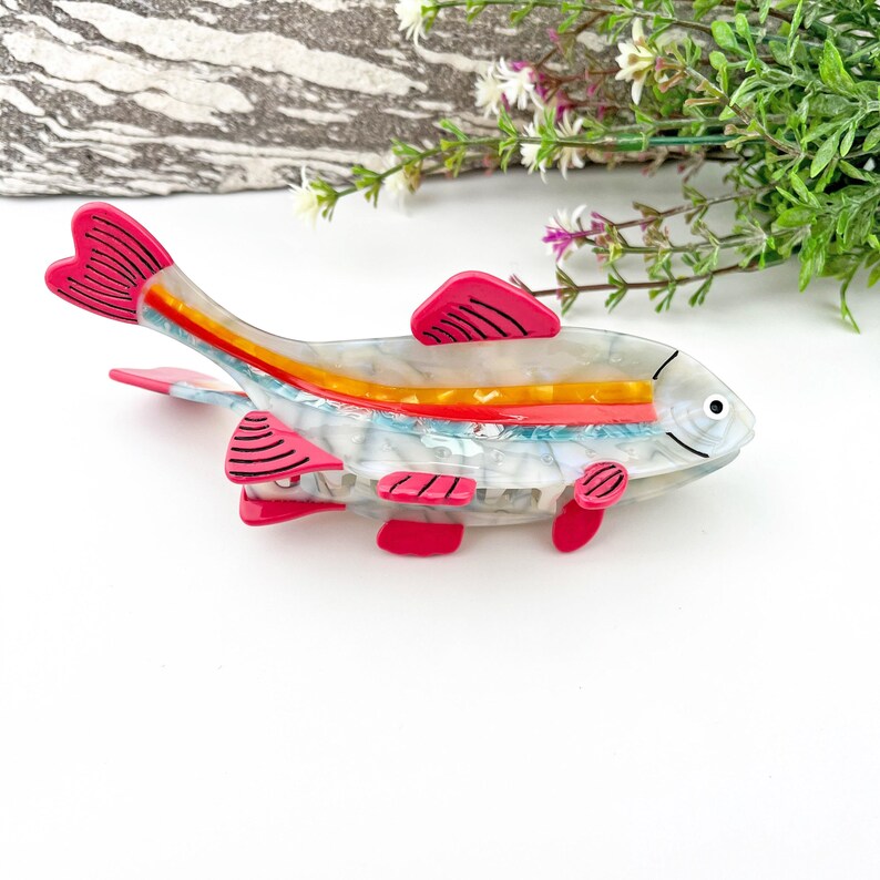 May include: A white and blue fish-shaped hair clip with pink accents. The fish has a red, orange, and blue stripe down its back.