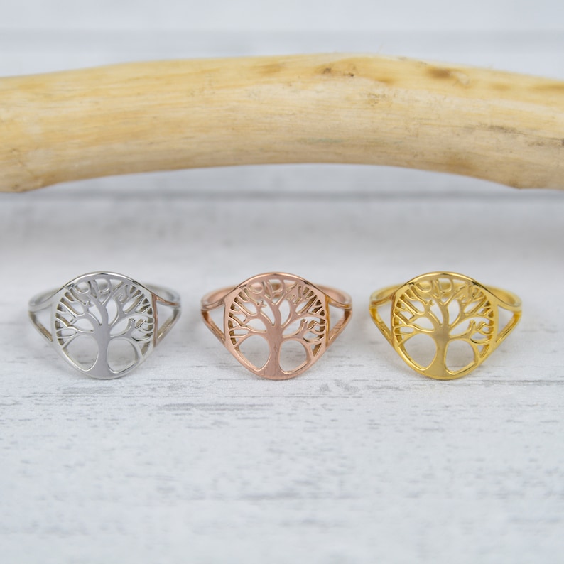 May include: Three rings with a tree of life design. The rings are silver, rose gold, and gold. The tree of life is a popular symbol of growth and connection. The rings are set on a white wooden surface.