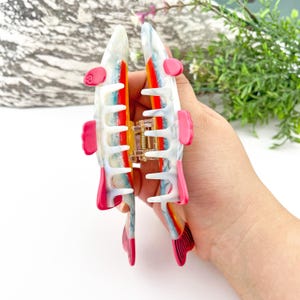 May include: A large white and blue hair claw with pink accents and a rainbow pattern. The claw is open and ready to hold hair.