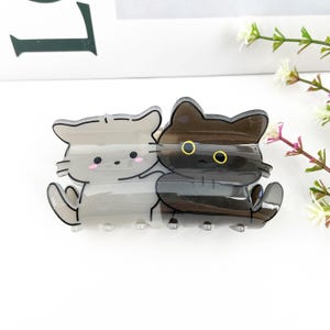 May include: A clear plastic hair clip featuring a design of two cartoon cats. The cat on the left is grey and the cat on the right is black. Both cats have black eyes with yellow pupils.