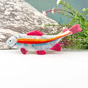 May include: A colorful, abstract fish-shaped hair clip with a white, blue, orange, and red design. The clip has a pink outline and is made of plastic.