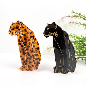 May include: Two plastic hair clips shaped like leopards. One clip is brown with black spots and the other is black. Both clips have gold outlines.