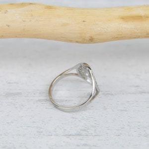 May include: A silver-colored ring with a tree-of-life design. The ring has a delicate, openwork pattern and a slender band. The ring is set against a white wooden surface.
