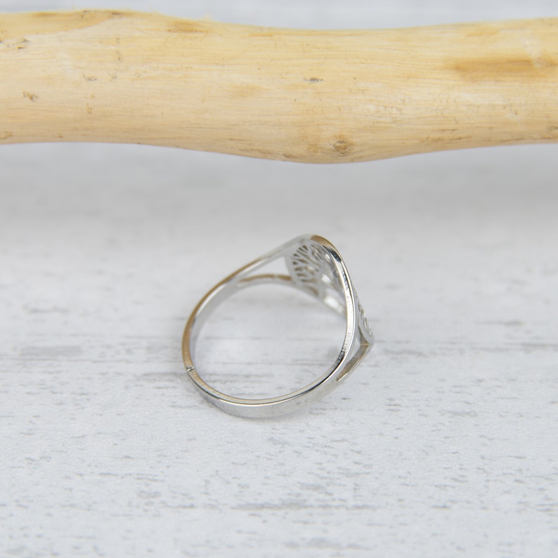 May include: A silver-colored ring with a tree-of-life design. The ring has a delicate, openwork pattern and a slender band. The ring is set against a white wooden surface.