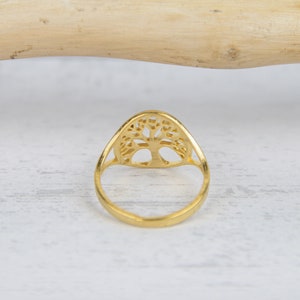 May include: A gold-colored ring featuring a tree of life design. The ring has a smooth, polished band and a detailed, openwork tree motif. The ring is set against a light-colored, textured surface.
