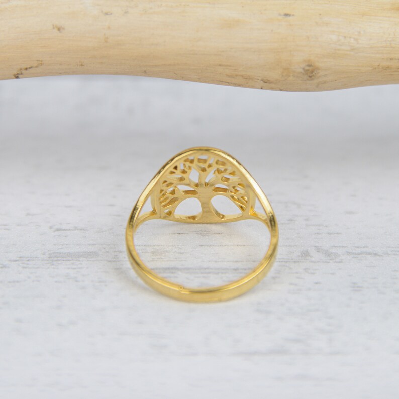 May include: A gold-colored ring featuring a tree of life design. The ring has a smooth, polished band and a detailed, openwork tree motif. The ring is set against a light-colored, textured surface.
