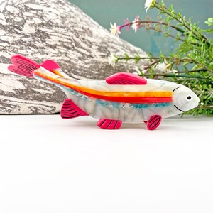May include: A white, pink, and orange fish brooch with a striped pattern. The fish has a pink tail and fins.
