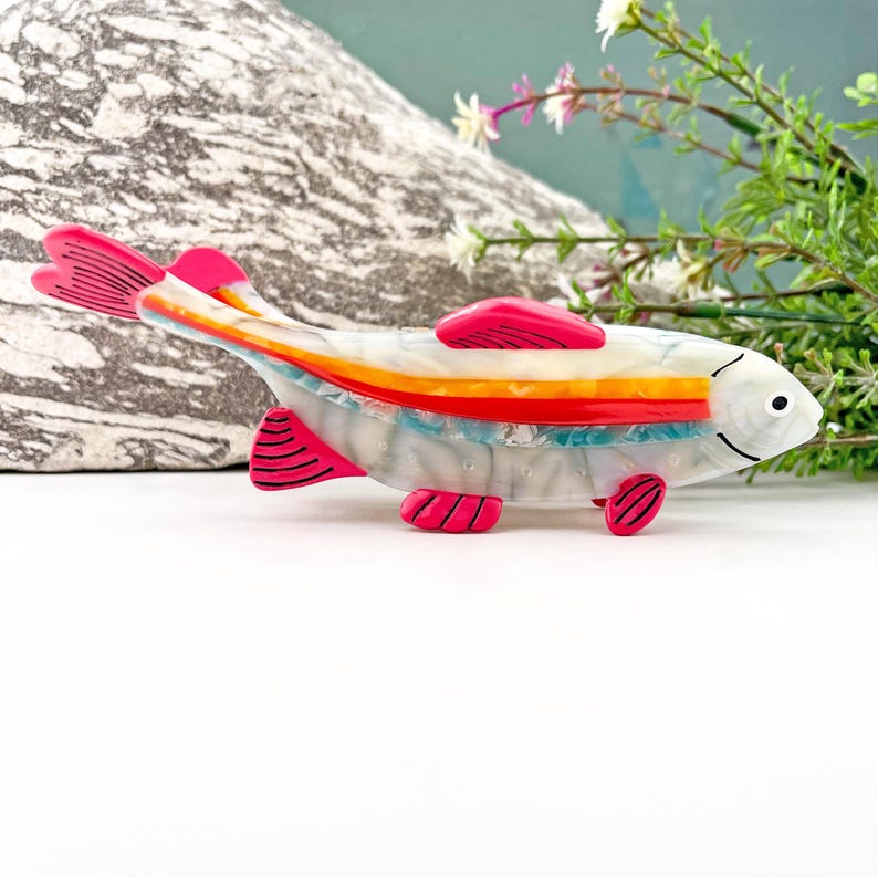 May include: A white, pink, and orange fish brooch with a striped pattern. The fish has a pink tail and fins.