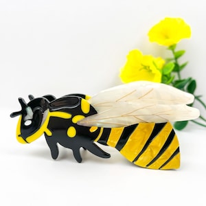 May include: A bee-shaped brooch with a black and yellow body and translucent wings. The brooch has a detailed design with a glossy finish. The bee is set against a white background with yellow flowers.