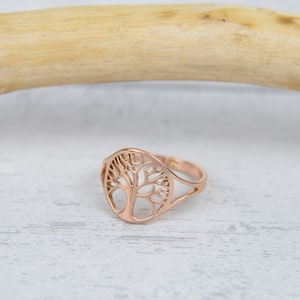 May include: A rose gold ring featuring a cut-out tree design. The ring has a delicate, openwork pattern of branches and a circular frame. The ring is set against a white and light gray background with a wooden stick.
