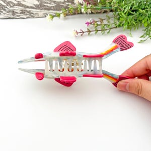 May include: A white and pink plastic hair claw shaped like a fish with a rainbow design. The claw has a gold metal spring.