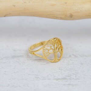 May include: A gold-colored ring featuring a cut-out tree design. The ring has a smooth, polished finish and a delicate, openwork pattern. The tree design is centered on the ring's face, with a slender band.