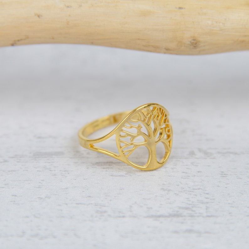 May include: A gold-colored ring featuring a cut-out tree design. The ring has a smooth, polished finish and a delicate, openwork pattern. The tree design is centered on the ring's face, with a slender band.