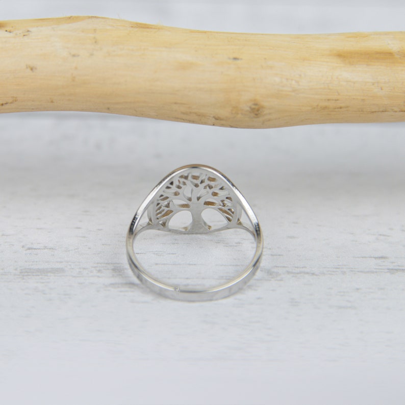 May include: A silver ring featuring a tree of life design. The ring has a polished finish and a detailed cutout of a tree within an oval shape. The ring is set against a white wooden surface.