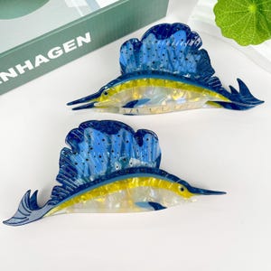 May include: Two blue and yellow marlin fish hair clips. The fish have a blue and white patterned fin and a yellow body with a white iridescent sheen.