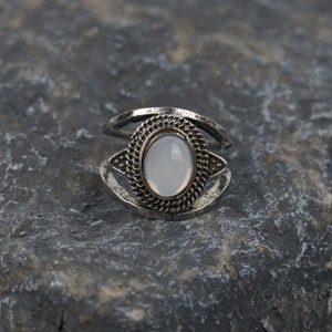 May include: A silver ring featuring an oval white stone set within an ornate frame. The ring has a double band design.