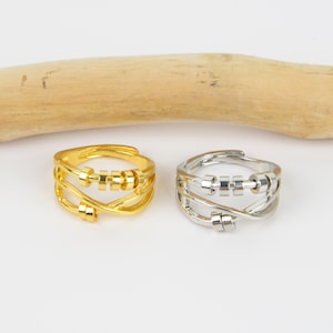 May include: Two adjustable rings, one gold and one silver, featuring a fidget bead design. The rings have a crisscross band and a row of small, movable beads. They are displayed on a white surface with a wooden element in the background.