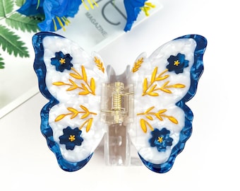 May include: Butterfly-shaped hair clip with a white and blue marbled design. The clip features gold-colored leaf and flower accents, and a clear plastic clasp. The clip is a decorative hair accessory.