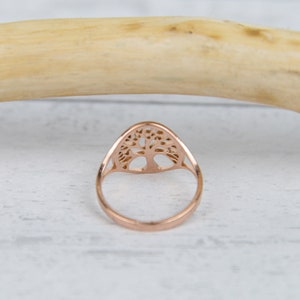 May include: Rose gold ring featuring a tree of life design. The ring has a delicate, openwork pattern of a tree with branches and roots. The ring is set against a white and light wood background.