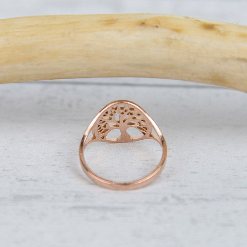 May include: Rose gold ring featuring a tree of life design. The ring has a delicate, openwork pattern of a tree with branches and roots. The ring is set against a white and light wood background.
