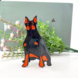 May include: Black and orange enamel pin of a Doberman Pinscher dog sitting with its legs crossed. The pin has a glossy finish.