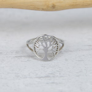 May include: A silver-colored ring featuring a tree of life design. The ring has a detailed cutout of a tree with branches and a trunk. The ring is set against a white wooden surface.