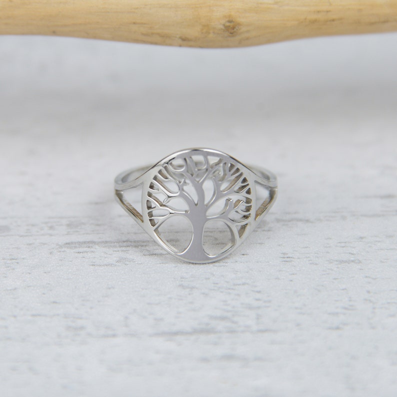 May include: A silver-colored ring featuring a tree of life design. The ring has a detailed cutout of a tree with branches and a trunk. The ring is set against a white wooden surface.