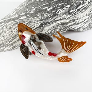 Puede incluir: Un broche de resina blanca, roja y marrón con forma de pez koi. El pez tiene un cuerpo blanco con marcas rojas y negras y una aleta caudal marrón.